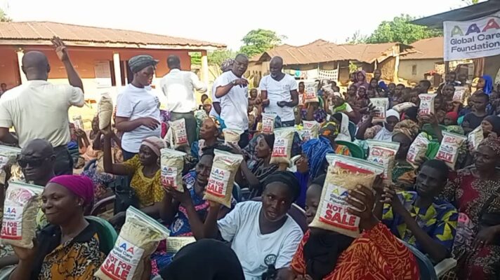 The scene depicts a community gathering, possibly during a charity event where aid is being distributed. Multiple people are seated in plastic chairs, and many of them are holding small packets or boxes that have "NOT FOR SALE" printed on them, indicating that these items are donated and not to be sold. The crowd consists of a mix of individuals, predominantly adults, dressed in various colorful and patterned clothing, which suggests a cultural or traditional context, likely African. Volunteers or staff members are visible among the seated individuals, standing as they distribute the packets. The expressions on people's faces range from neutral to smiles, suggesting a positive atmosphere. The setting appears to be outdoors, in a communal space, perhaps a courtyard or open area of a village or town. The building structures in the background are simple and residential in style. Overall, the image conveys a sense of community engagement and support.